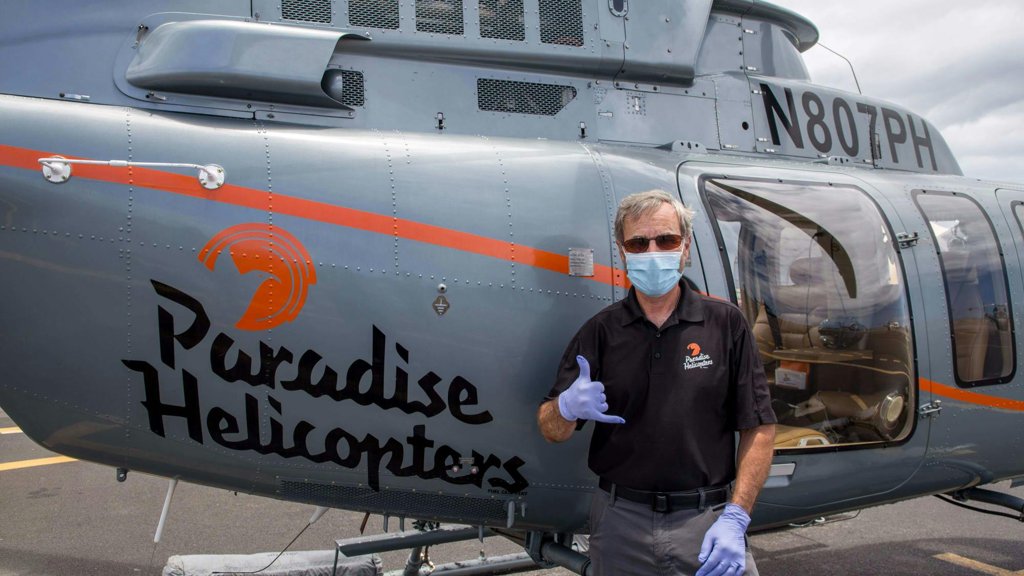 man doing the "hang loose" hand gesture wearing latex gloves and a surgical mask in front of a helicopter